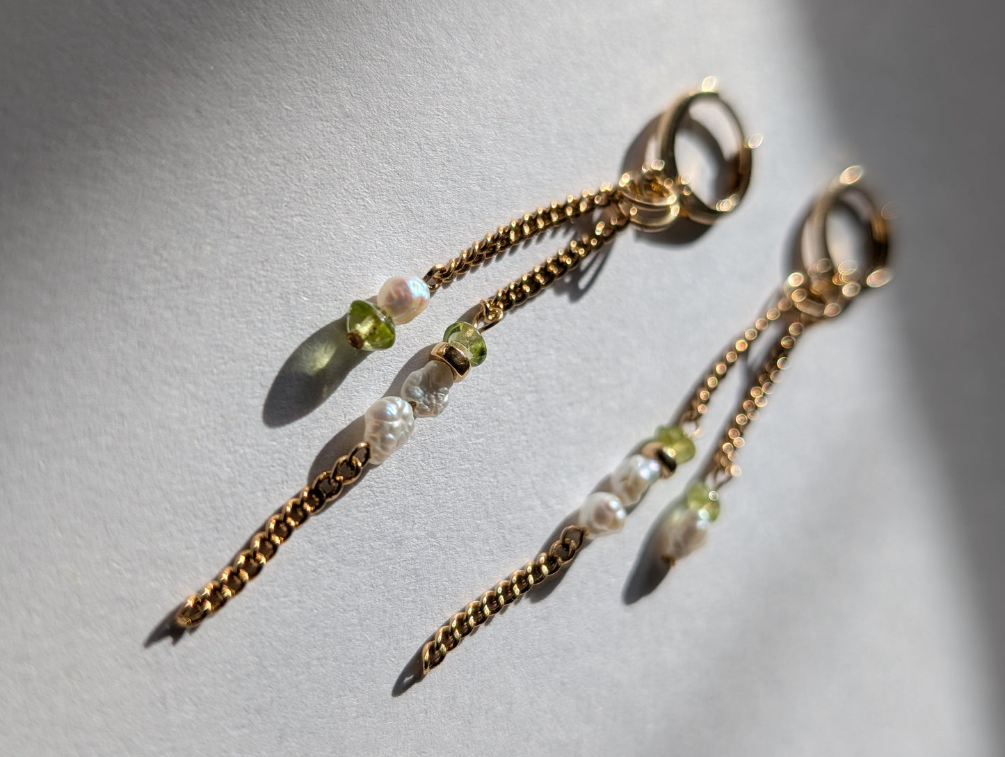A pair of dangling earrings with 14K gold-filled hoops, chains, and beads made of genuine Freshwater Pearls and bright green Peridot stones, displayed against a light background.